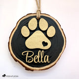 Close-up of a personalized black-painted wood slice dog ornament featuring a layered gold paw print with heart cutout and the name “Bella” in matching gold script.