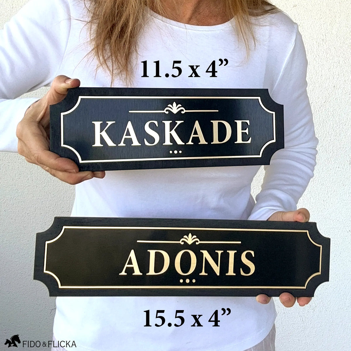 Person holding two black and gold layered wood horse stall name plates for size comparison, showing an 11×4 inch sign reading ‘Kaskade’ above a 15×4 inch extended-length sign reading ‘ADONIS'.