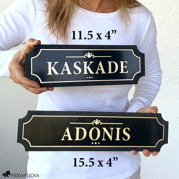 Person holding two black and gold layered wood horse stall name plates for size comparison, showing an 11×4 inch sign reading ‘Kaskade’ above a 15×4 inch extended-length sign reading ‘ADONIS'.