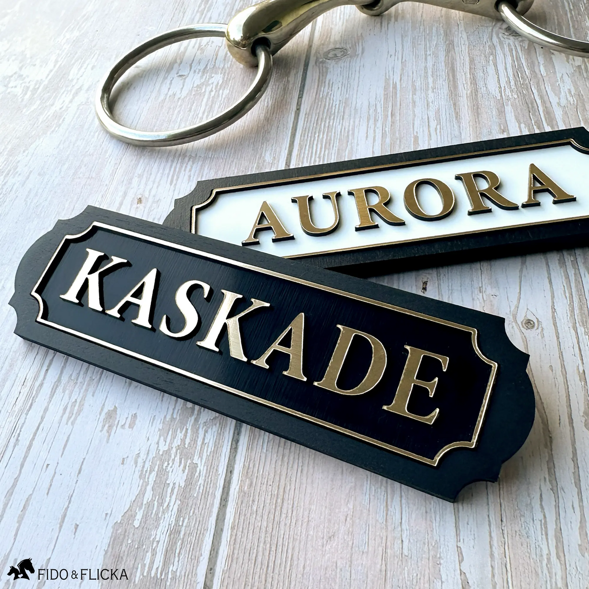 Two personalized acrylic and wood horse nameplates with brushed gold lettering and scalloped borders, reading "Aurora" on a white plate and "Kaskade" on a black plate, styled on weathered wood beside a brass snaffle bit.