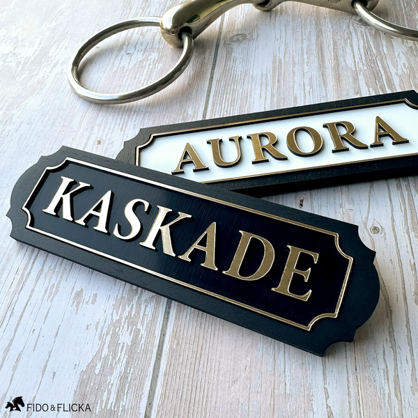 Two personalized acrylic and wood horse nameplates with brushed gold lettering and scalloped borders, reading "Aurora" on a white plate and "Kaskade" on a black plate, styled on weathered wood beside a brass snaffle bit.
