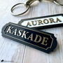 Two personalized acrylic and wood horse nameplates with brushed gold lettering and scalloped borders, reading "Aurora" on a white plate and "Kaskade" on a black plate, styled on weathered wood beside a brass snaffle bit.