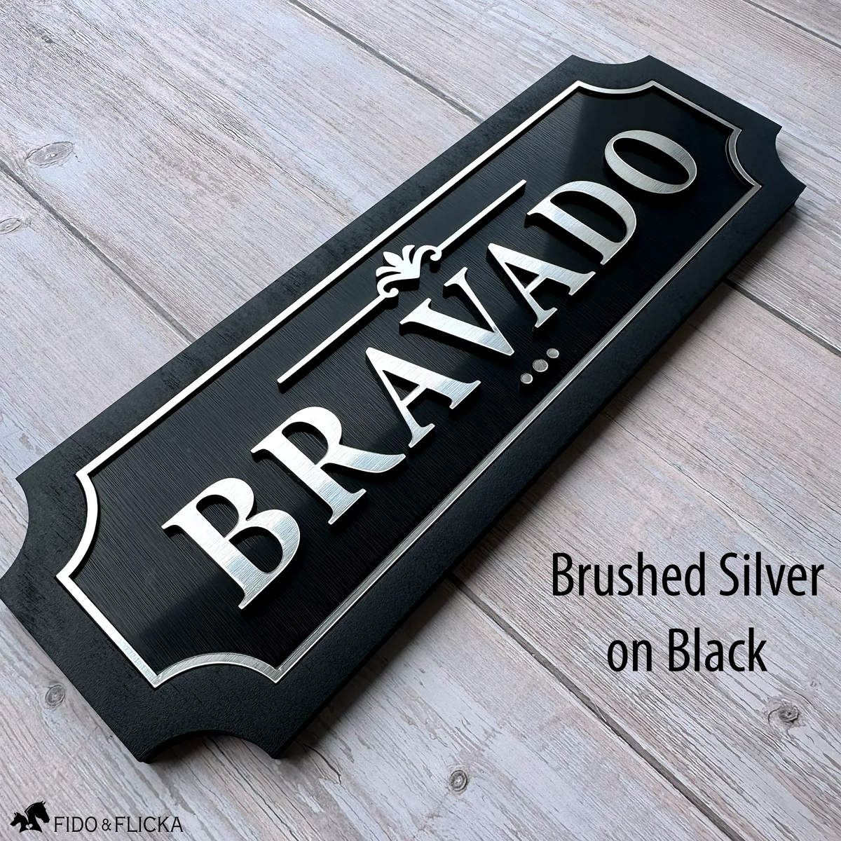 Personalized horse stall sign with raised brushed silver acrylic letters spelling Bravado on brushed black acrylic base with sealed black wood frame displayed on weathered wood surface by Fido and Flicka