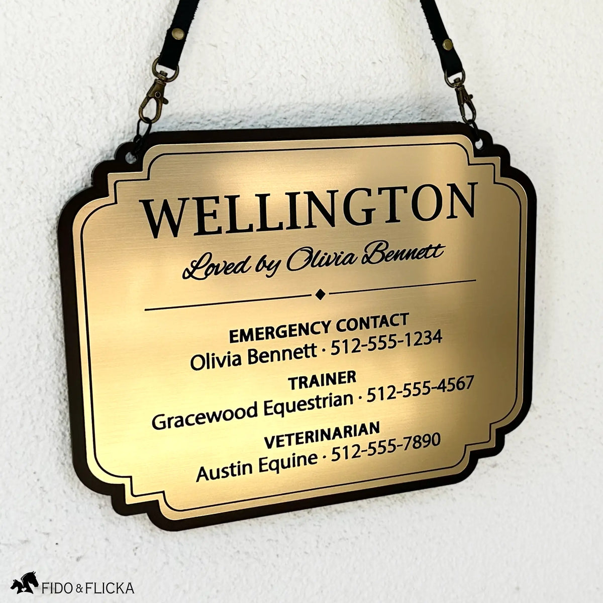 Brushed gold acrylic stall card sign for Wellington hanging from black straps against a white wall, with a decorative black border and emergency, trainer, and veterinarian contact details.