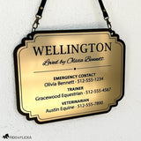 Brushed gold acrylic stall card sign for Wellington hanging from black straps against a white wall, with a decorative black border and emergency, trainer, and veterinarian contact details.