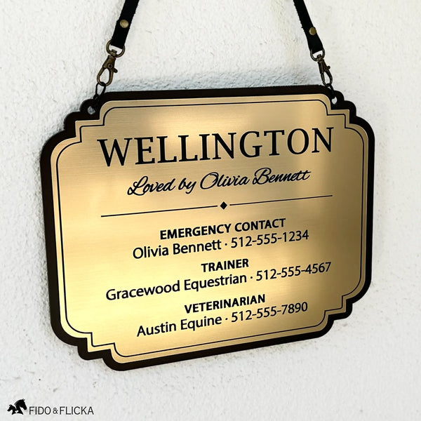 Brushed gold acrylic stall card sign for Wellington hanging from black straps against a white wall, with a decorative black border and emergency, trainer, and veterinarian contact details.
