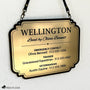 Brushed gold acrylic stall card sign for Wellington hanging from black straps against a white wall, with a decorative black border and emergency, trainer, and veterinarian contact details.