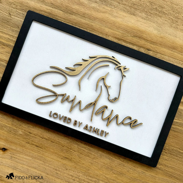 Custom horse stall nameplate by Fido & Flicka featuring a gold horse silhouette and the name “Sundance” with the inscription “Loved by Ashley.” The design is laser-cut on a white wood background with a black frame, blending modern elegance and equestrian artistry.