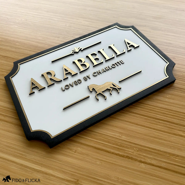 Personalized horse stall plaque 11.5 by 7.5 inches with raised brushed gold acrylic letters spelling Arabella loved by Charlotte with trot horse icon on white acrylic base panel and sealed black wood frame by Fido and Flicka