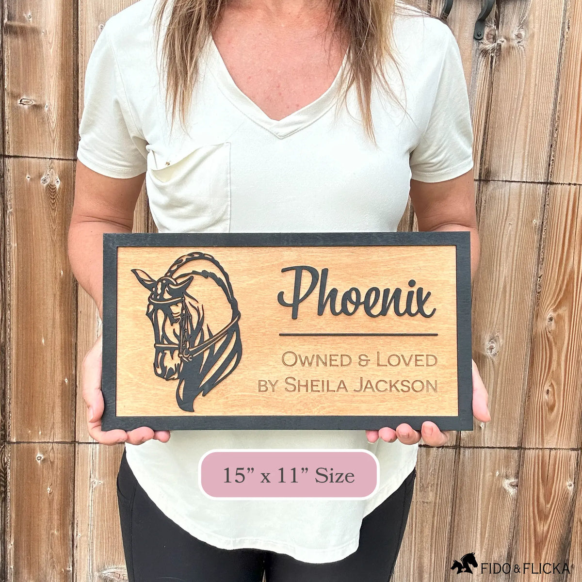 Woman holding custom wooden horse stall sign made from Baltic birch. Personalized plaque shows black dressage horse head design with engraved text horse name, Owned & Loved by owner. Sign measures 15” x 11”, rustic barn name plate.