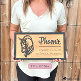 Woman holding custom wooden horse stall sign made from Baltic birch. Personalized plaque shows black dressage horse head design with engraved text horse name, Owned & Loved by owner. Sign measures 15” x 11”, rustic barn name plate.