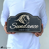 Person holding a personalized horse stall sign featuring layered horse artwork, custom horse name, and engraved text on a black acrylic plaque to show the sign's size and scale.