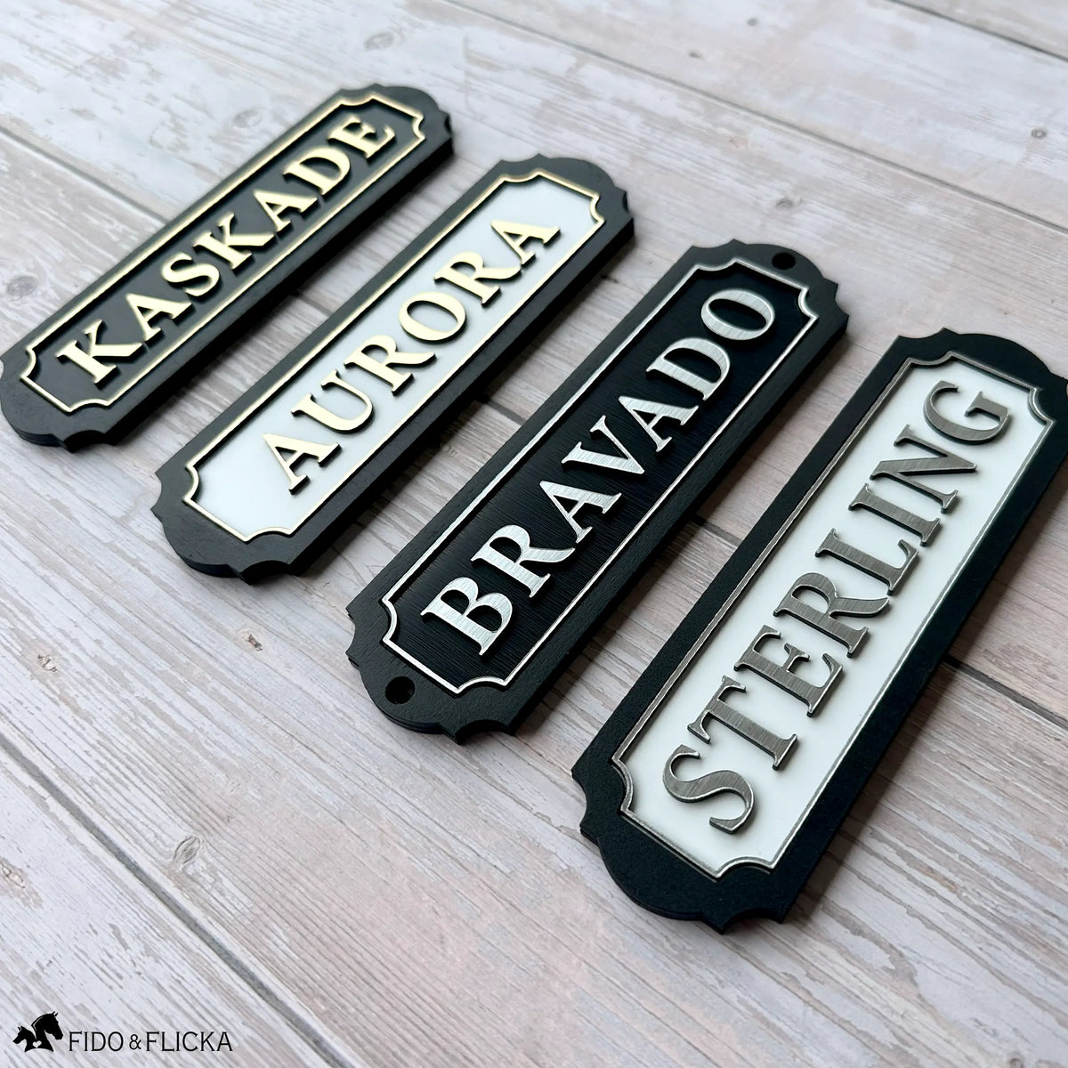Four tack room nameplates shown side by side in the available finishes: brushed gold lettering on black, brushed gold on white, brushed silver on black, and brushed silver on white, each on a black wood backer with scalloped edges.