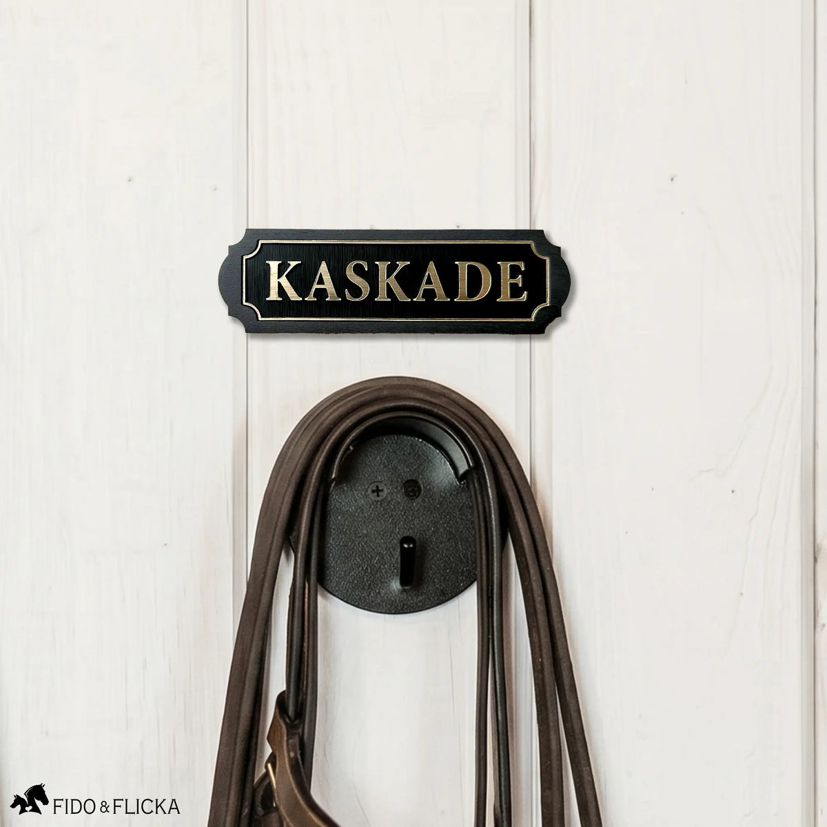 Black acrylic and wood tack room nameplate with brushed gold lettering reading "Kaskade," mounted on a white barn wall above a bridle hook holding a brown leather bridle.