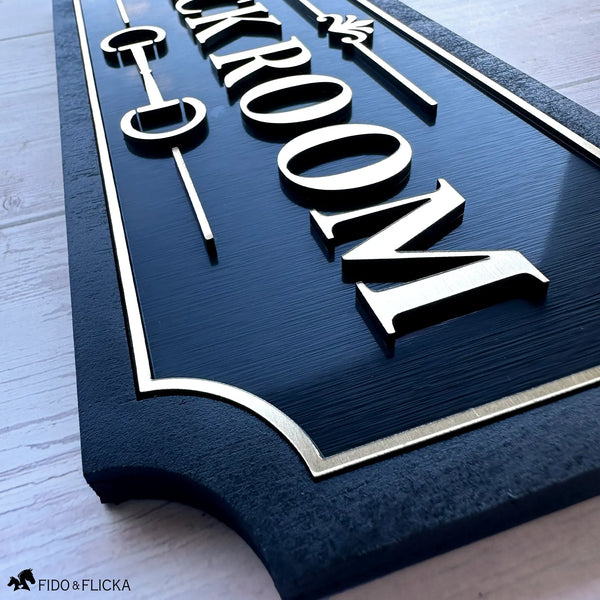 Close-up of a custom Tack Room sign showing dimensional brushed gold lettering, layered acrylic details, and black wood backing.