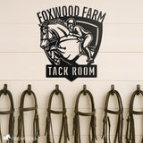 Custom metal tack room sign featuring a show jumping horse and rider with personalized text ‘Foxwood Farm Tack Room,’ mounted above bridle hooks in an equestrian barn