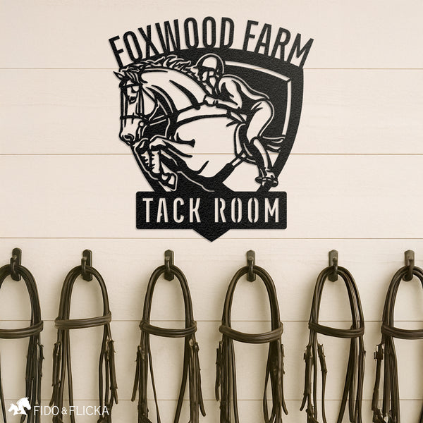 Custom metal tack room sign featuring a show jumping horse and rider with personalized text ‘Foxwood Farm Tack Room,’ mounted above bridle hooks in an equestrian barn
