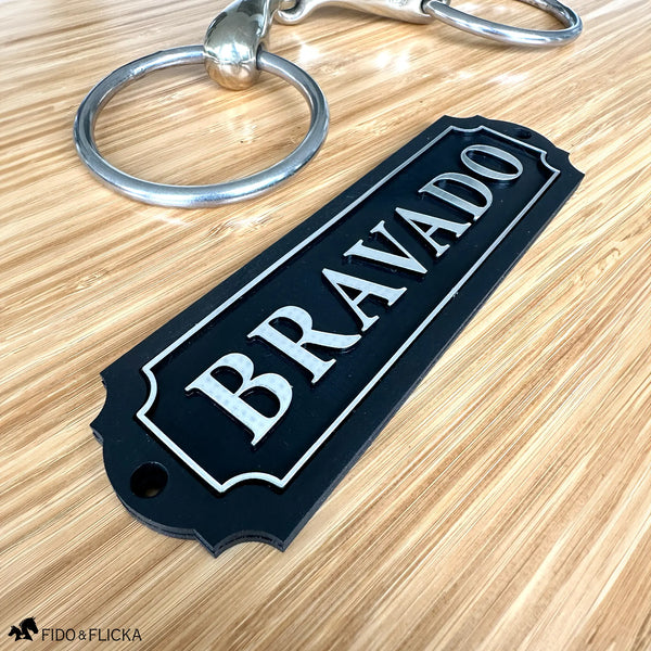 Personalized black acrylic and wood tack room nameplate with raised brushed silver lettering reading "Bravado," a brushed silver border, scalloped edges, and two mounting holes, shown on light wood beside a silver snaffle bit.