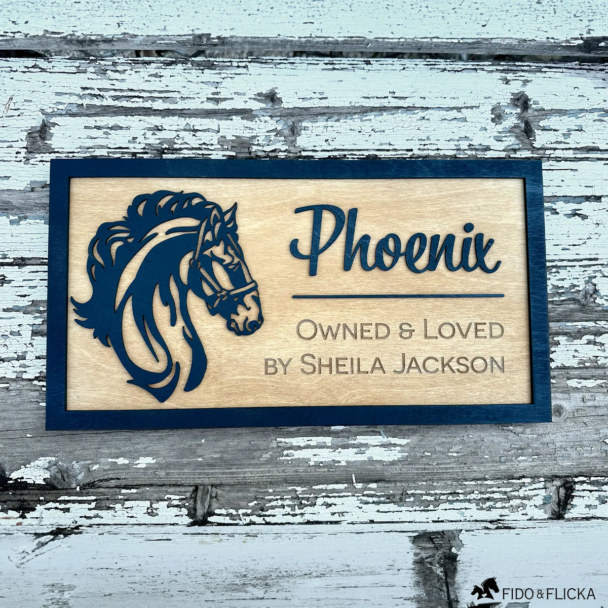 Custom wooden horse stall sign made from Baltic birch. Features layered black horse head silhouette and engraved text “Phoenix, Owned & Loved by Sheila Jackson.” Personalized barn name plate displayed on rustic weathered wood background