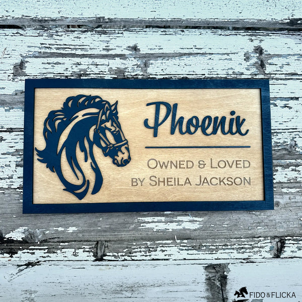 Custom wooden horse stall sign made from Baltic birch. Features layered black horse head silhouette and engraved text “Phoenix, Owned & Loved by Sheila Jackson.” Personalized barn name plate displayed on rustic weathered wood background