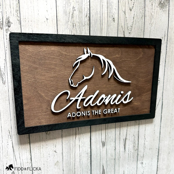 Close-up angled view of a custom horse stall nameplate by Fido & Flicka featuring a white horse silhouette and the name “Adonis” with the inscription “Adonis the Great.” The design is mounted on a dark walnut wood background with a black frame, showcasing elegant craftsmanship and modern equestrian style.