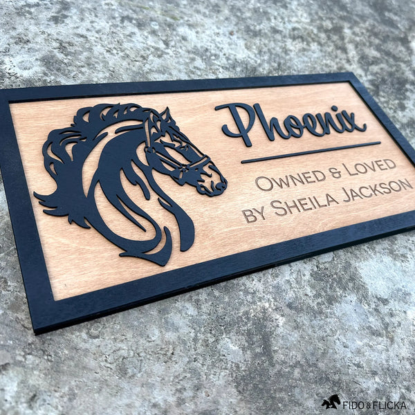 Angled view of custom wooden horse stall sign made from Baltic birch. Layered black horse head silhouette with engraved text “Phoenix, Owned & Loved by Sheila Jackson.” Personalized barn name plate shown on stone background